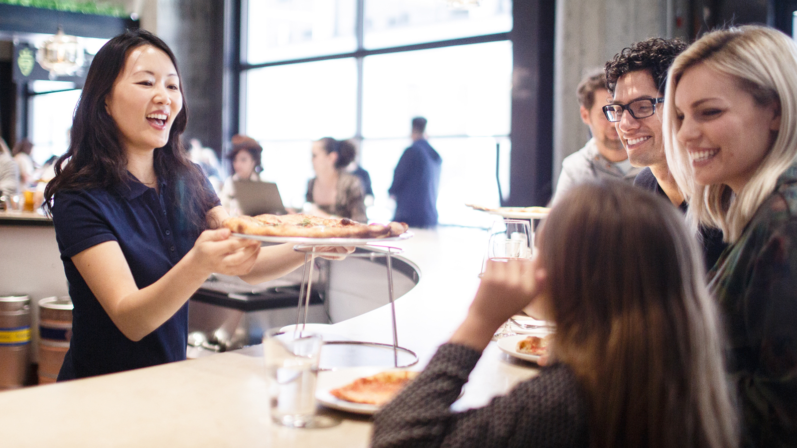 Gambar seorang wanita pemilik restaurant yang menawarkan pizza kepada pelanggan dan menggunakan fasilitas biaya merchant dari Visa